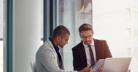 Two men are sitting at a table in a modern office. One is wearing a light-colored blazer and taking notes, while the other is in a dark suit holding a tablet. A laptop is open on the table, and they seem to be discussing something on the tablet.
